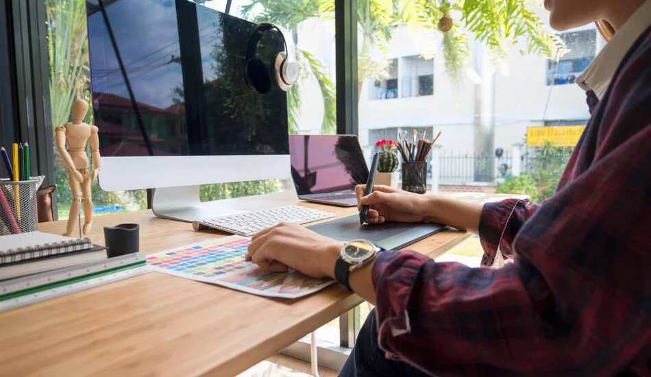 A designer working at a desk with a graphics tablet, color samples, and a large monitor, illustrating the manual effort involved in catalog design.